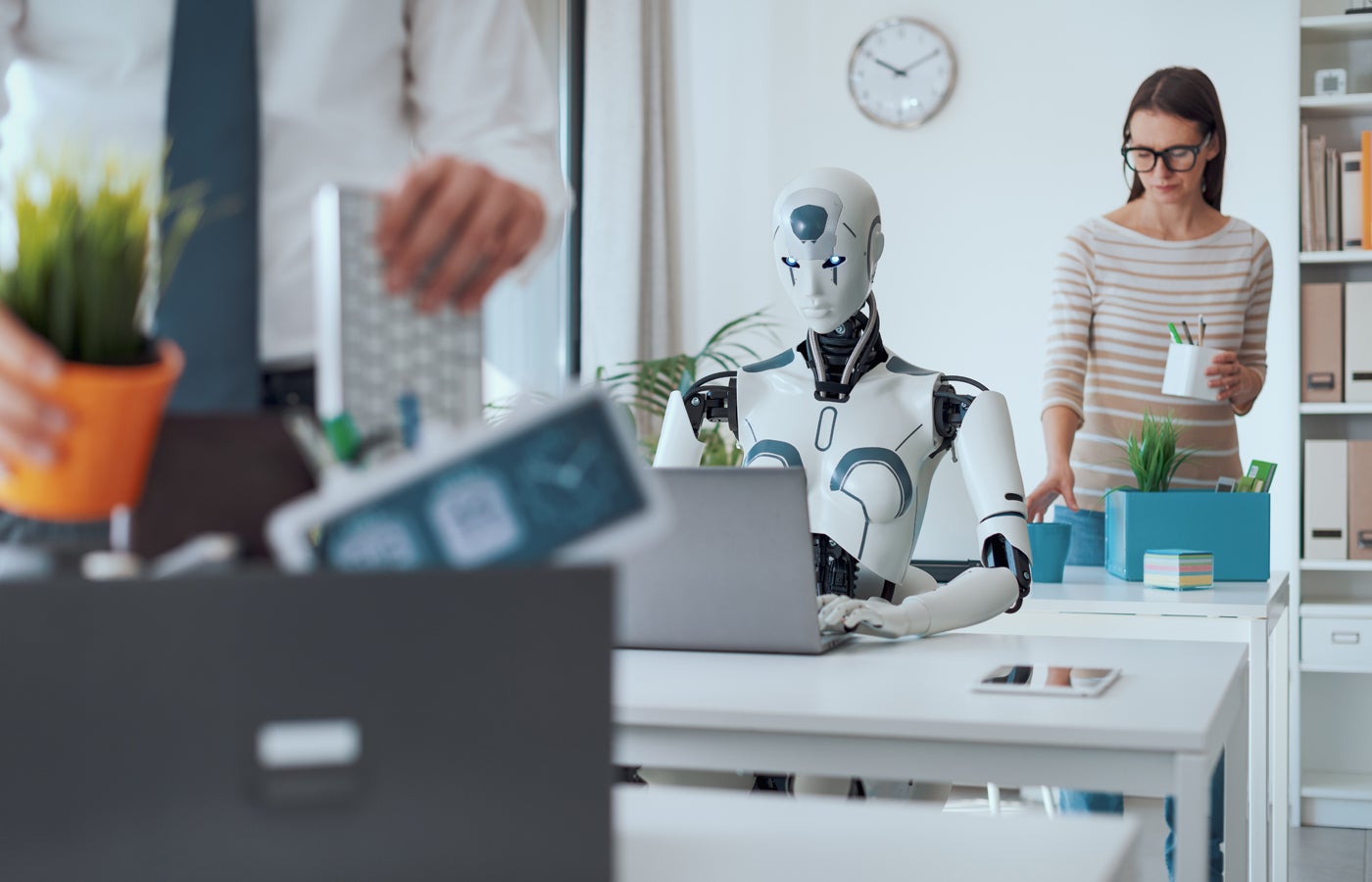 Dismissed business people packing their belongings and leaving the office, while a robot is sitting at the desk and working.