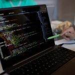 Focus on laptop screen with coded data and pen held by hand of young man.