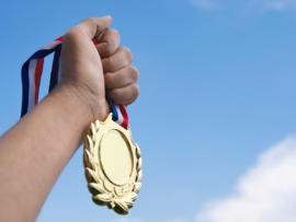 A hand triumphantly holds a gold medal aloft against a backdrop of clear blue sky.